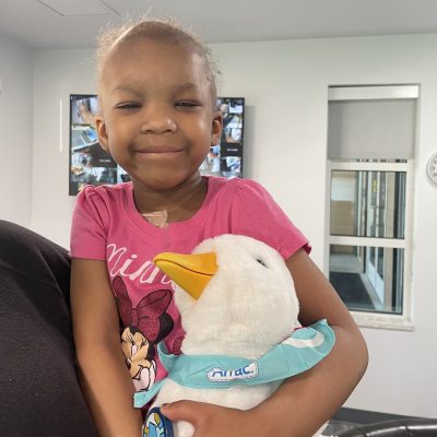 A young girl in a pink Minnie Mouse shirt smiles while hugging a white Aflac plush duck wearing a teal bandana.