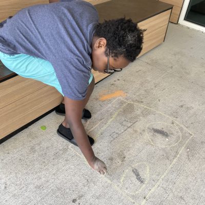 A young boy in a gray t-shirt and teal shorts leans over to draw a square with yellow and orange sidewalk chalk on a concrete patio.
