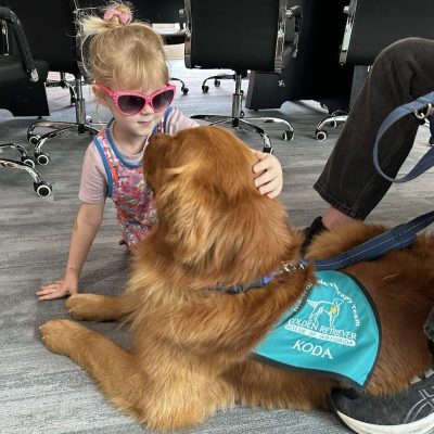 A young girl with blonde hair and pink sunglasses gently pets a golden retriever therapy dog named Koda. The dog wears a teal vest that reads "Hearts of Gold Therapy Team."