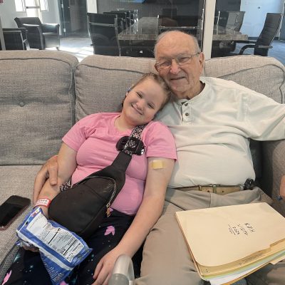 A dedicated older male volunteer with glasses, Mr. Joel, sits on a gray couch with his arm around a smiling young girl named Piper. Piper leans her head on his shoulder, and a file folder labeled "RMH Visits 2025" sits in Joel's lap.