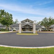 A wide shot of the modern, one-story Ronald McDonald House Charities of North Central Florida building. It features a prominent white gabled entryway, a paved circular driveway, and green landscaping under a bright blue sky.